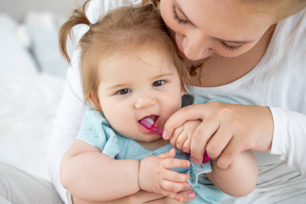 Mother brushing baby's first tooth with toothbrush while child laughing. Happy education of healthy lifestyle and baby care. Babyhood and parenthood.